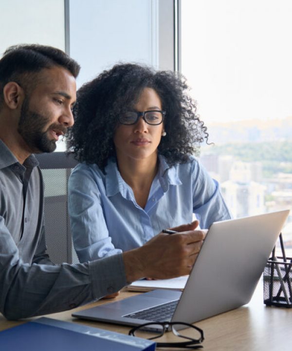 Indian male ceo executive manager mentor giving consultation on financial operations to female African American colleague intern using laptop sitting in modern office near panoramic window.